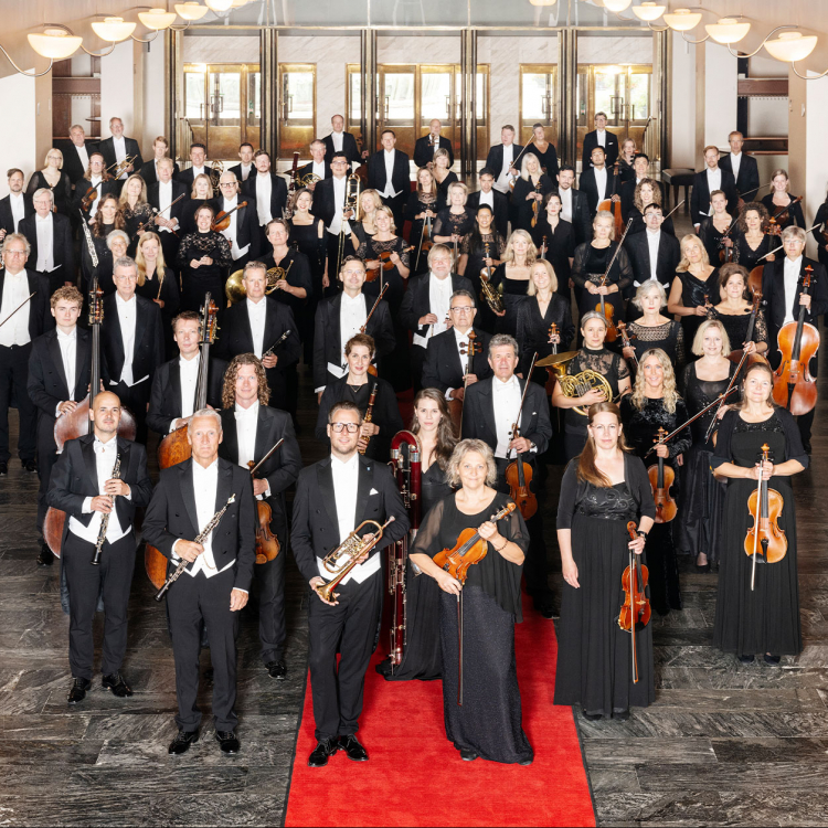 Orkesterbild där alla musiker är klädda i frack och långklänning och står på röda mattan i Konserthusets garderobshall. De tittar alla in i kameran.