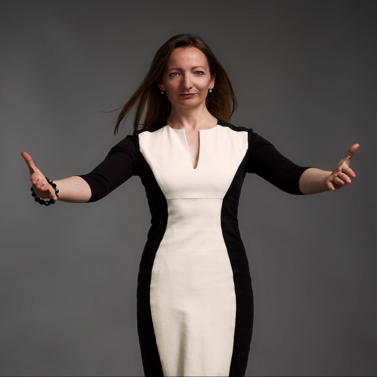 Katie Thomas står i svartvit klänning med händerna utsträkta mot kameran. Mot grå bakgrund.