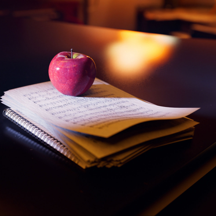 En hög med notblad ligger på en mörk flygel. Ovanpå ligger ett rött äpple och i bakgrunden lyser en gul lampa.
