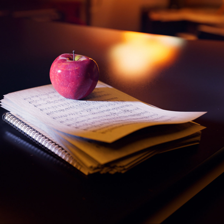 En hög med notblad ligger på en mörk flygel. Ovanpå ligger ett rött äpple och i bakgrunden lyser en gul lampa.