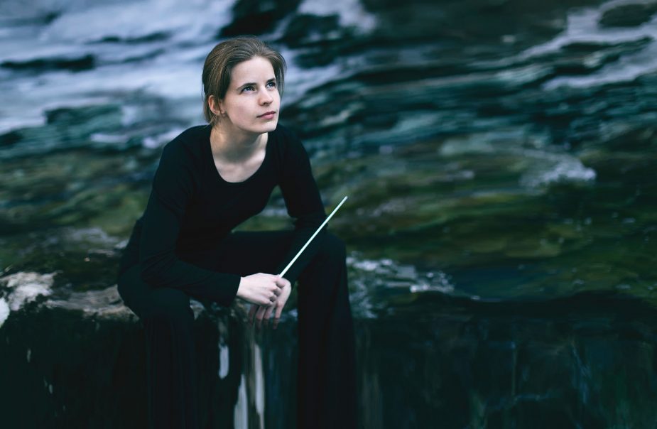 Kvinna sitter på sten vid vatten med dirigentpinne i handen.