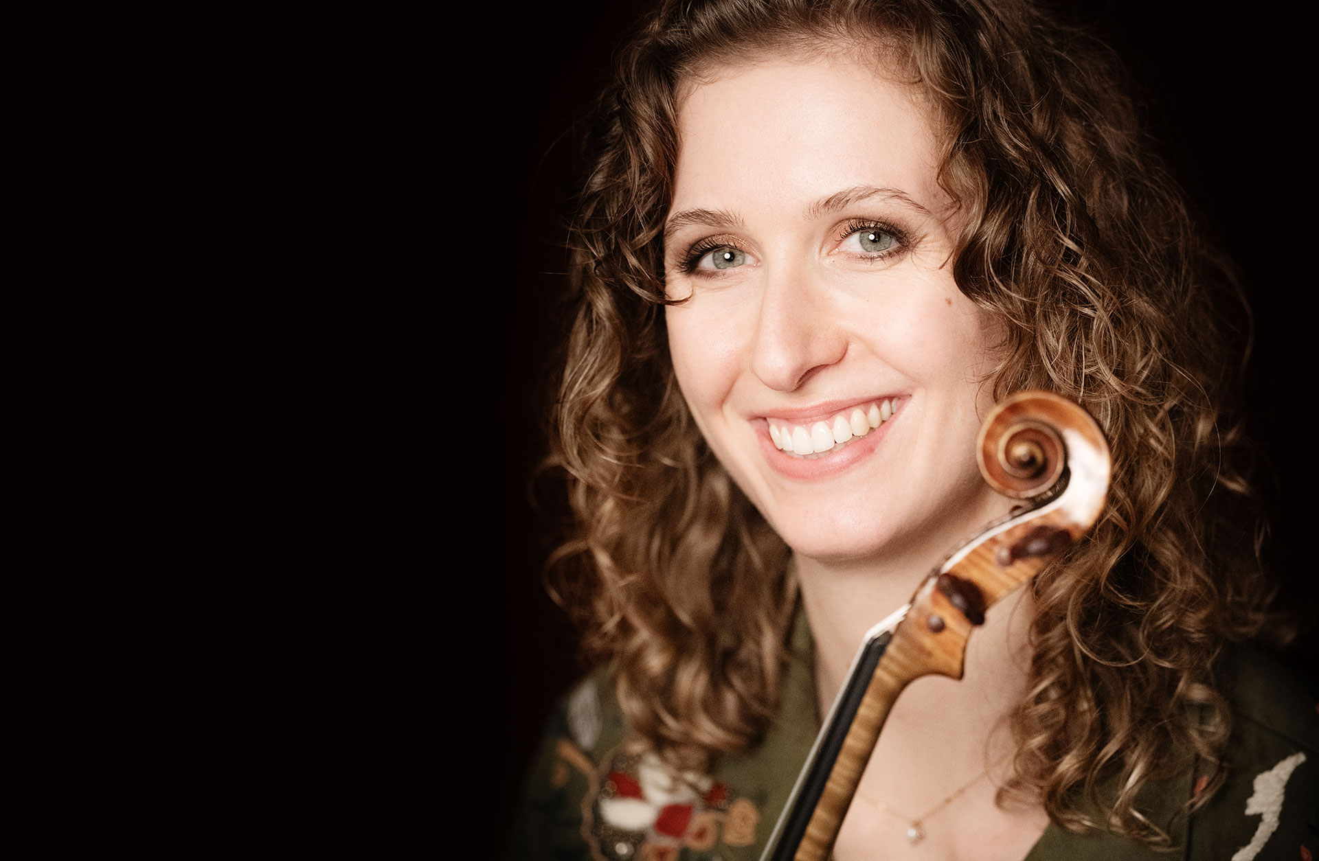 Porträtt av Clara Garde med sin fiol i famnen. Hon har lockigt långt cendréfärgat hår och ler mot kameran.