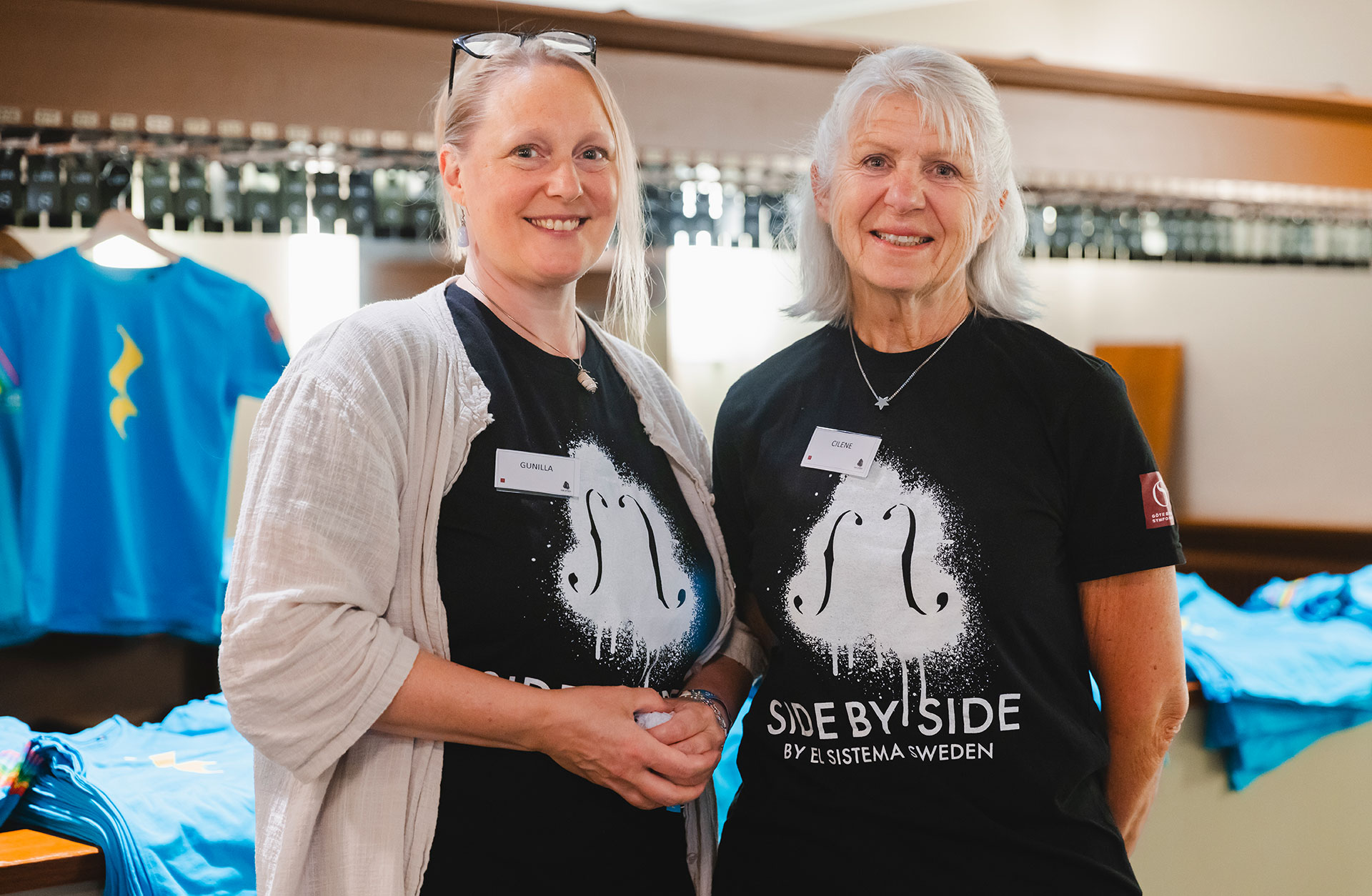 Två kvinnliga volontärer i svarta T-shirts med Side by Sides logga tittar in i kameran. De står i Konserthusets garderob.