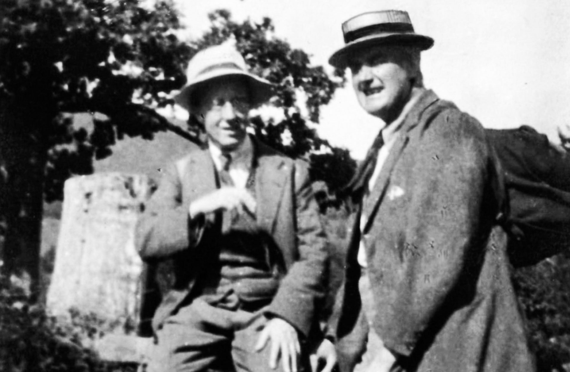 Arkivbild i svartvitt på två äldre herrar i kostym och hatt ute i naturen.