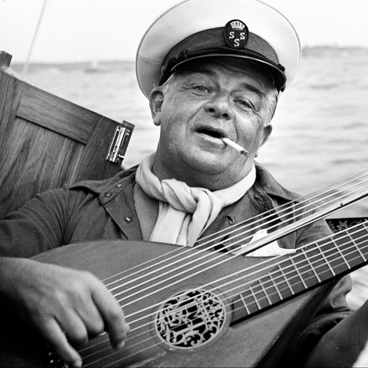 Bild på Evert Taube med instrument i handen ombord på båt med kaptensmössa.