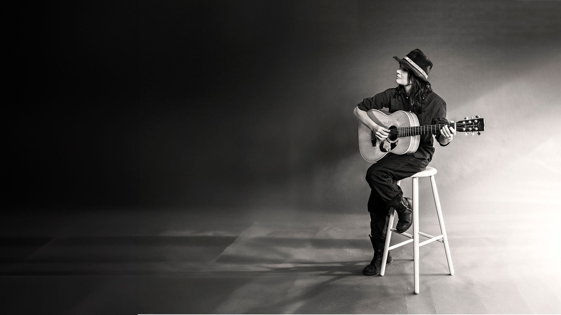 En kvinna sitter på en vit hög pall med en gitarr i knät. hon har en hatt på huvudet.