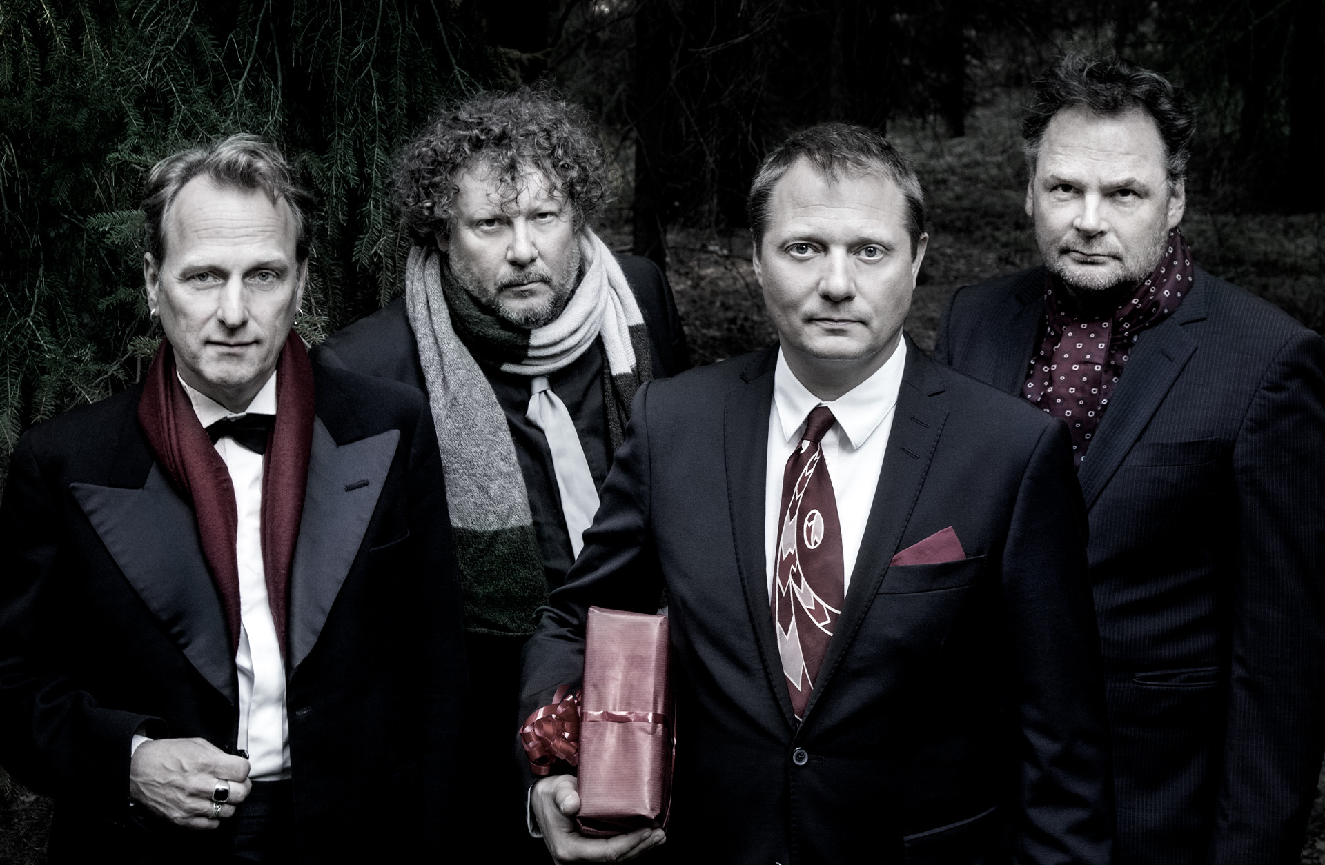 Fyra män i kostymer kollar in i kameran och mannen längst fram håller i ett rött paket