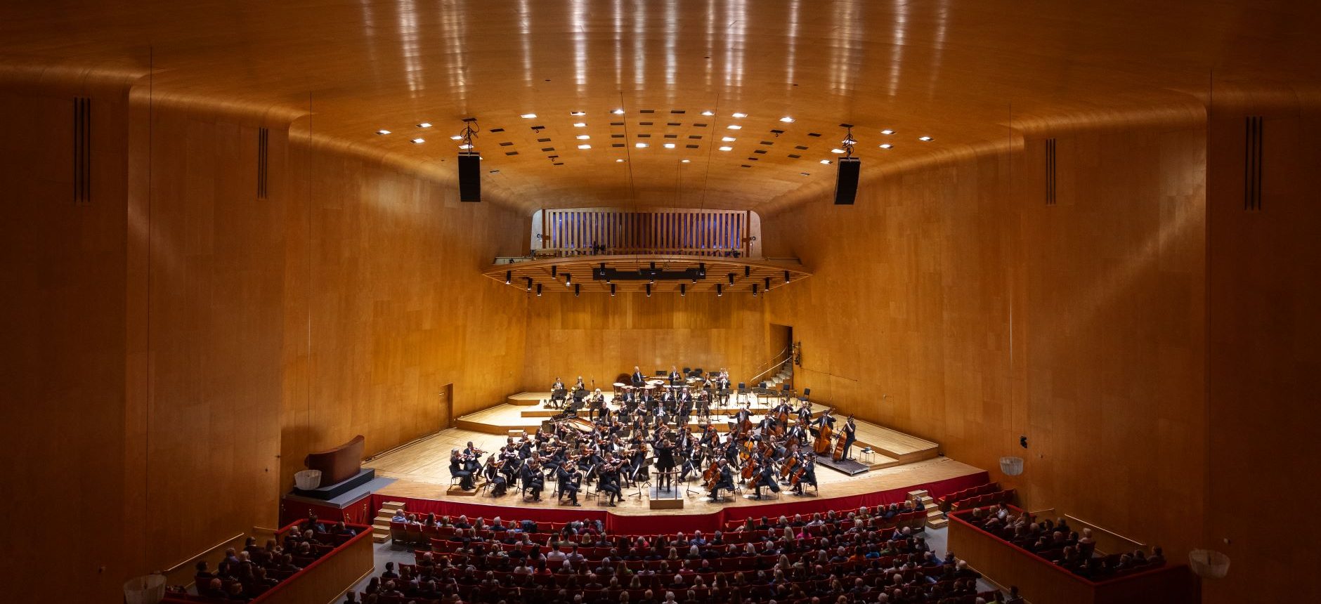 Utsikt över stora salen ovanifrån med publik sittandes ner, och full orkester på scenen.