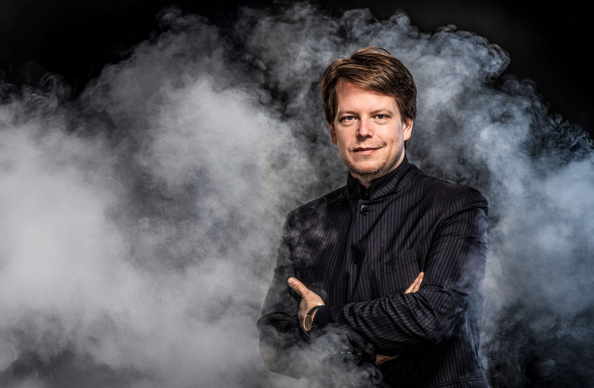 En man i randig mörk kavaj står i ett mörkt rum omgiven av rök.