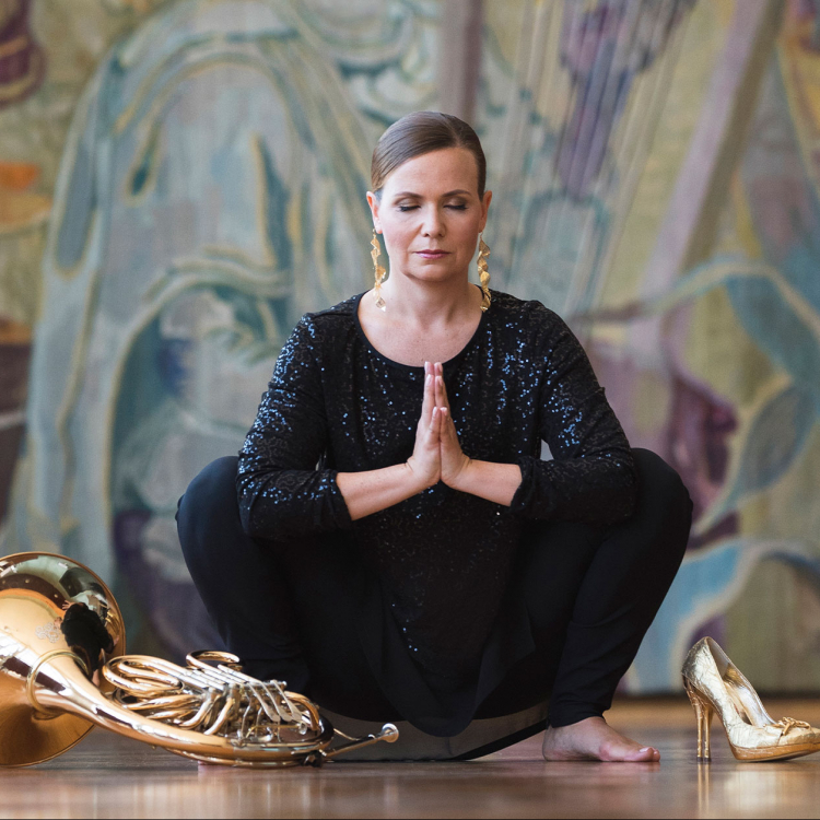 En finklädd sminkad kvinna sitter i en hukad yogaposition och blundar. Det ligger ett horn bredvid henne.