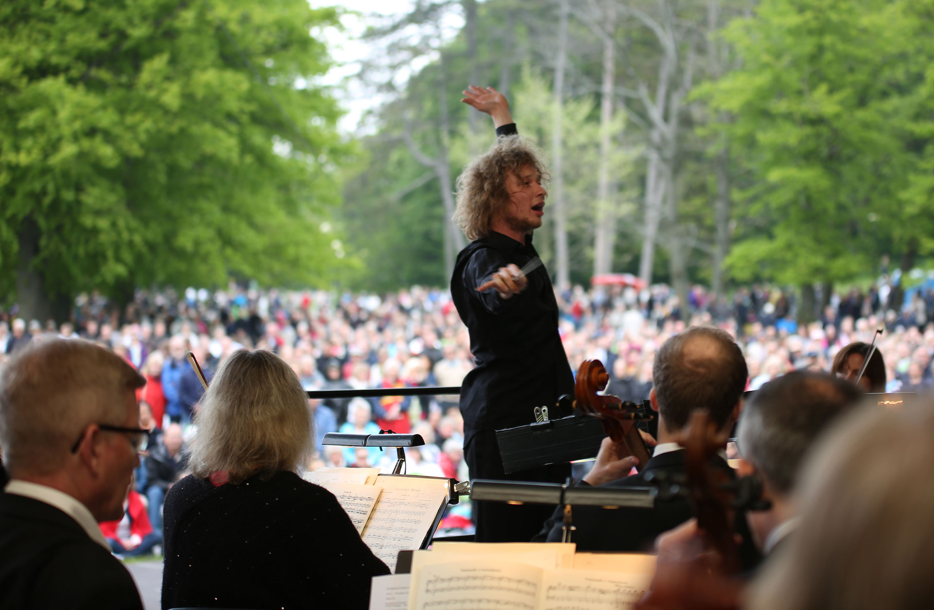 Santtu-Matias Rouvali dirigerar orkestern i Slottsskogen. Man ser ett stort folkhav bakom honom.