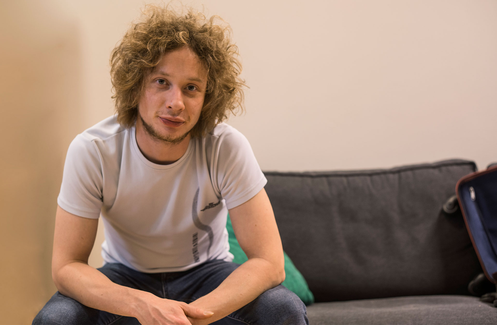 En ung man med burrigt blont hår sitter i en soffa. Han är klädd i T-shirt och blå jeans.