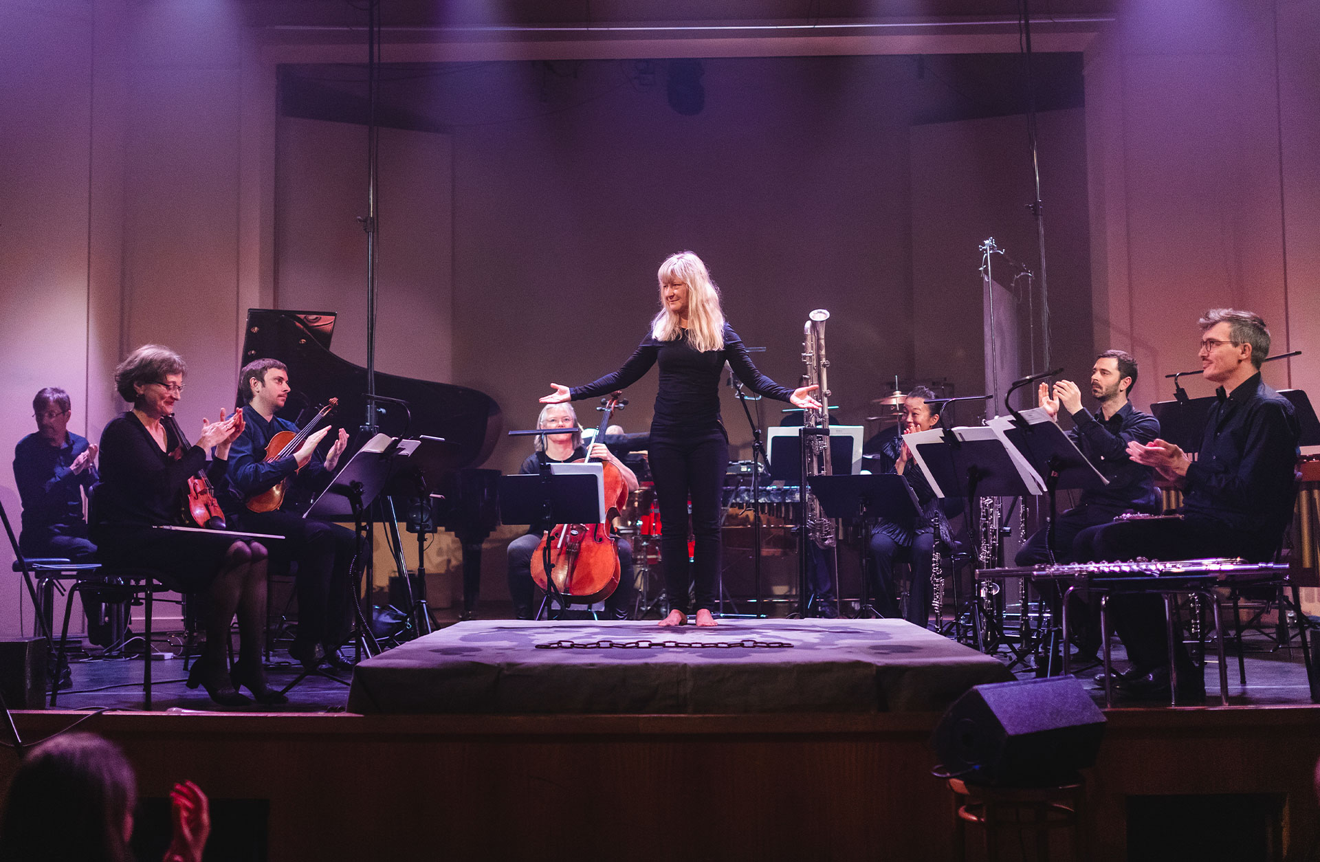 En svartklädd kvinna med långt blont hår står i strålkastarljus. Runt sig har hon en mindre orkester.