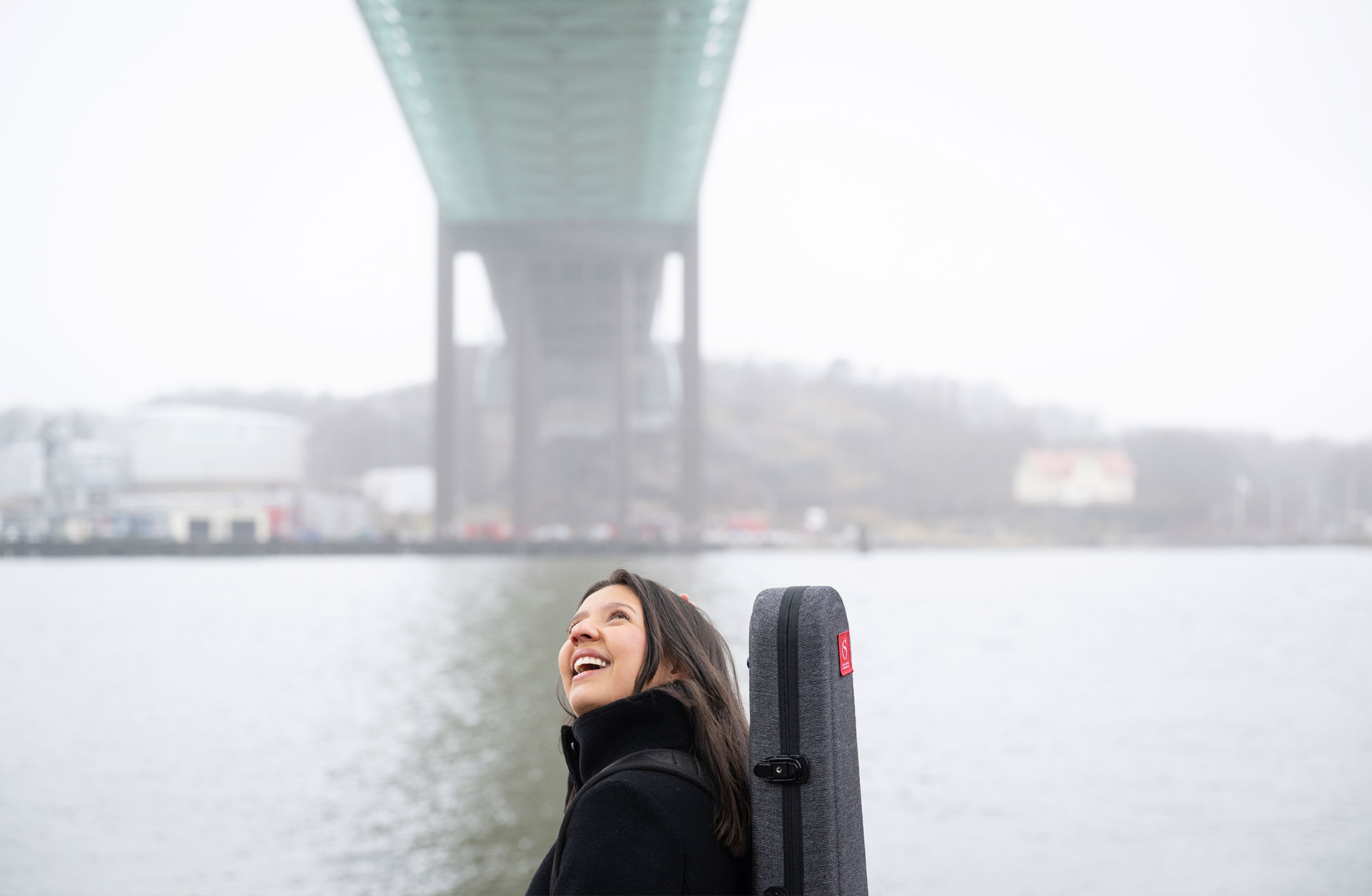 En ung kvinna går med sitt fiolcase ute. Hon står under en bro och tittar upp.