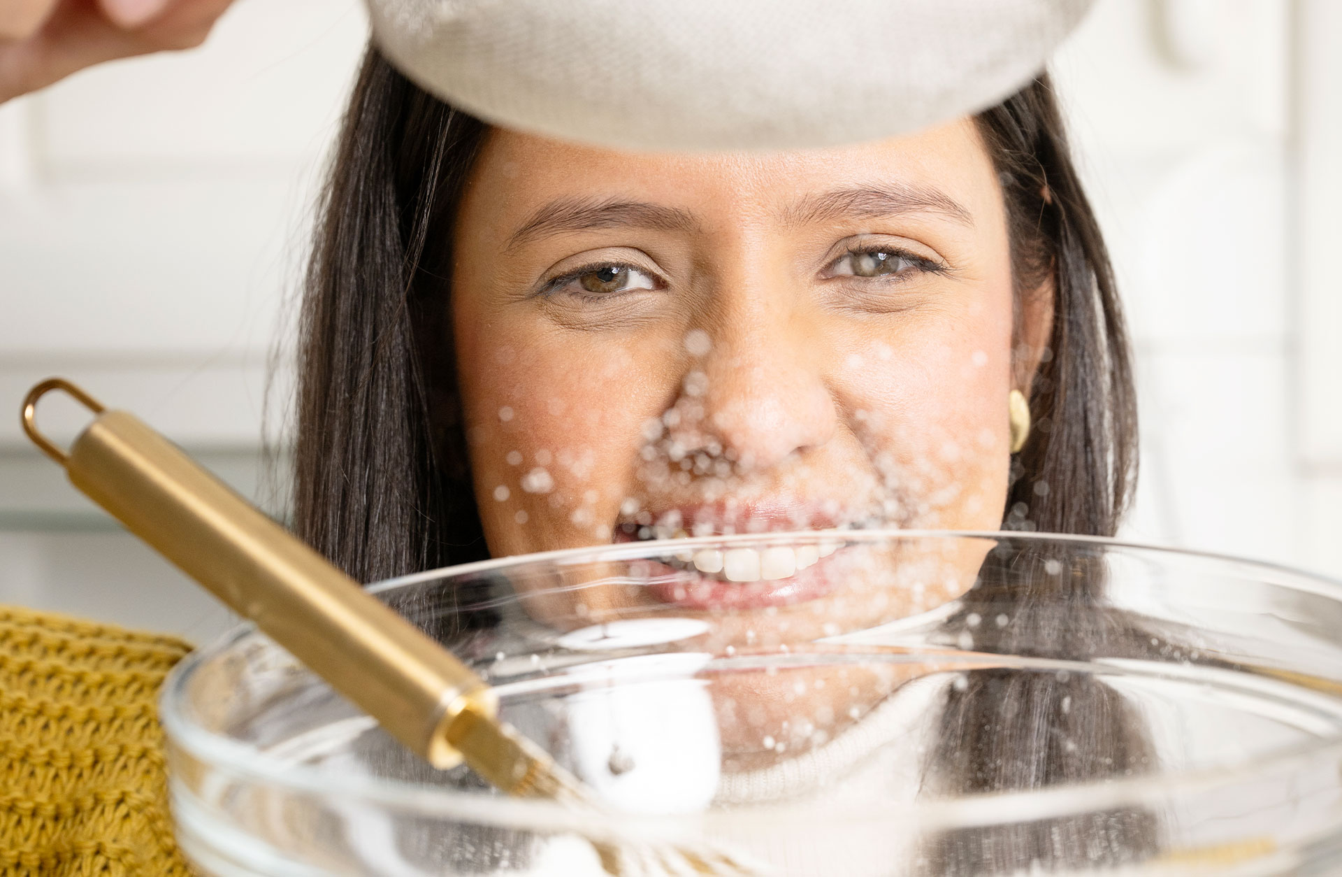 Ansiktet på en glad ung kvinna som silar mjöl i en bakskål framför ansiktet.