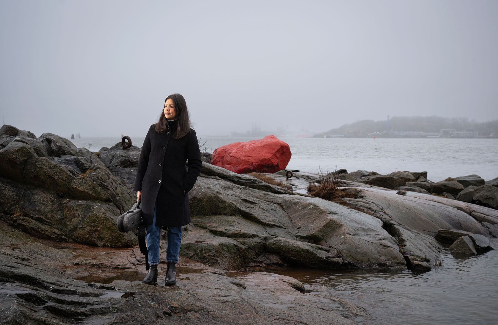 Emirzeth går med sitt fiolfodral på stranden vid Röda sten. Hon har en svart kappa på sig och man ser den röda stora stenen på en klippa bakom henne.