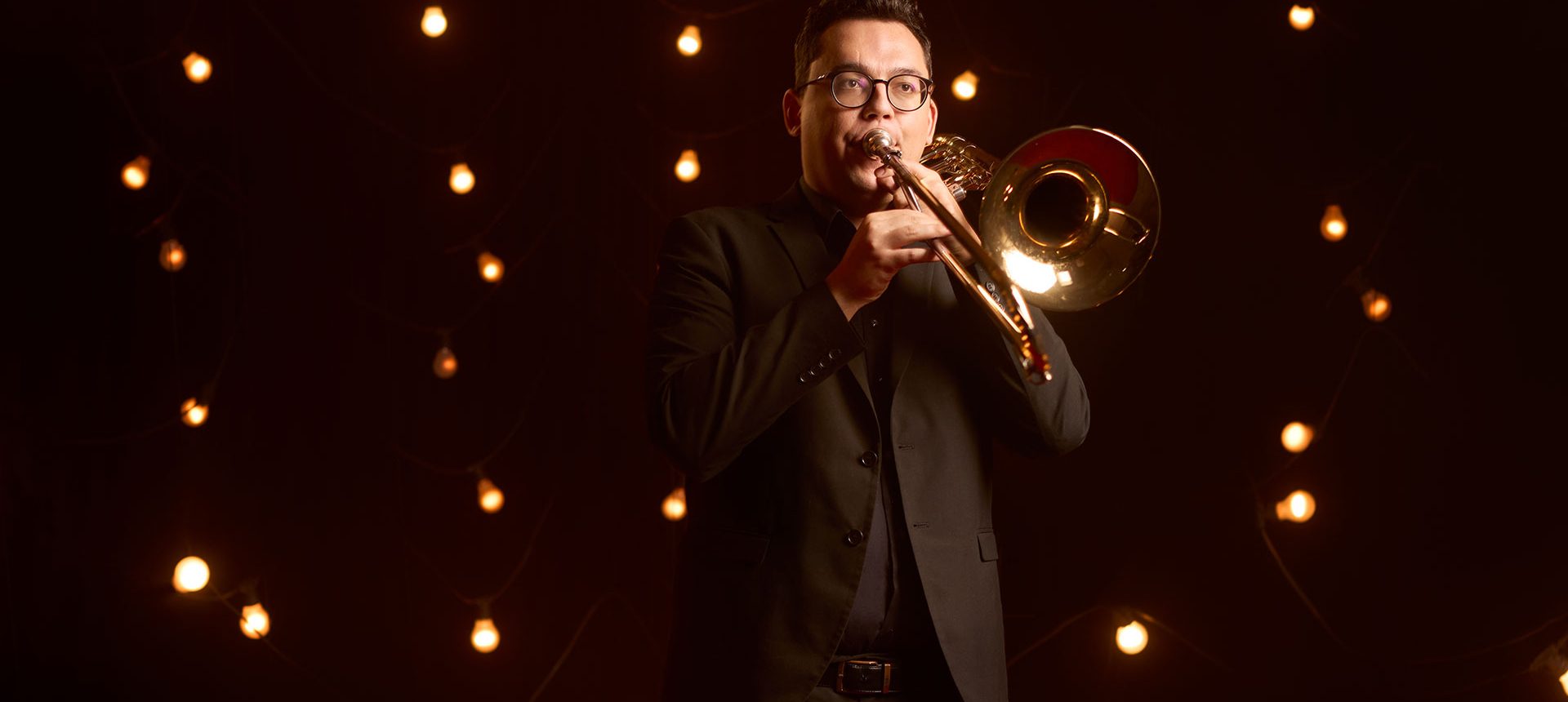 Man spelar trombon mot en mörk bakgrund med glödlampor.