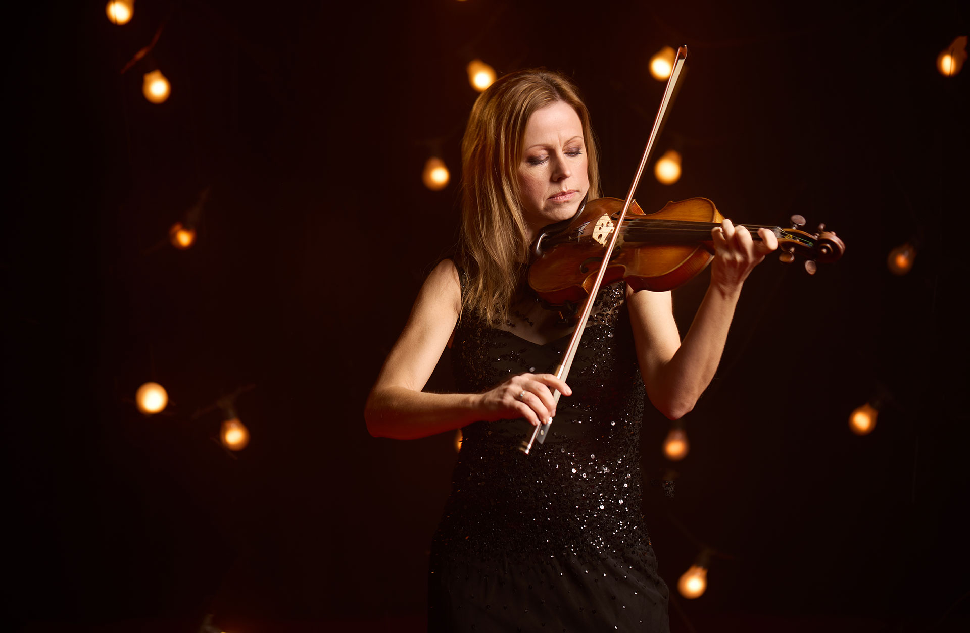 Kvinna spelar violin mot en bakgrund av svart och skimrande glödlampor.