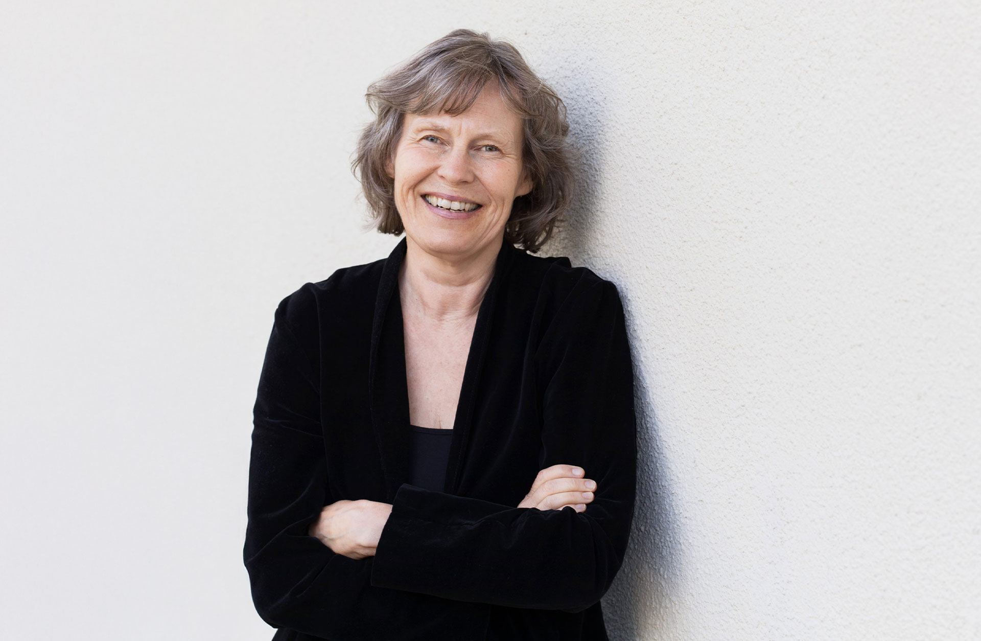 En glad kvinna med gråsprängt hår i kort frisyr står mot en vit vägg utomhus. hon har en svart kofta och håller armarna i kors över bröstet.