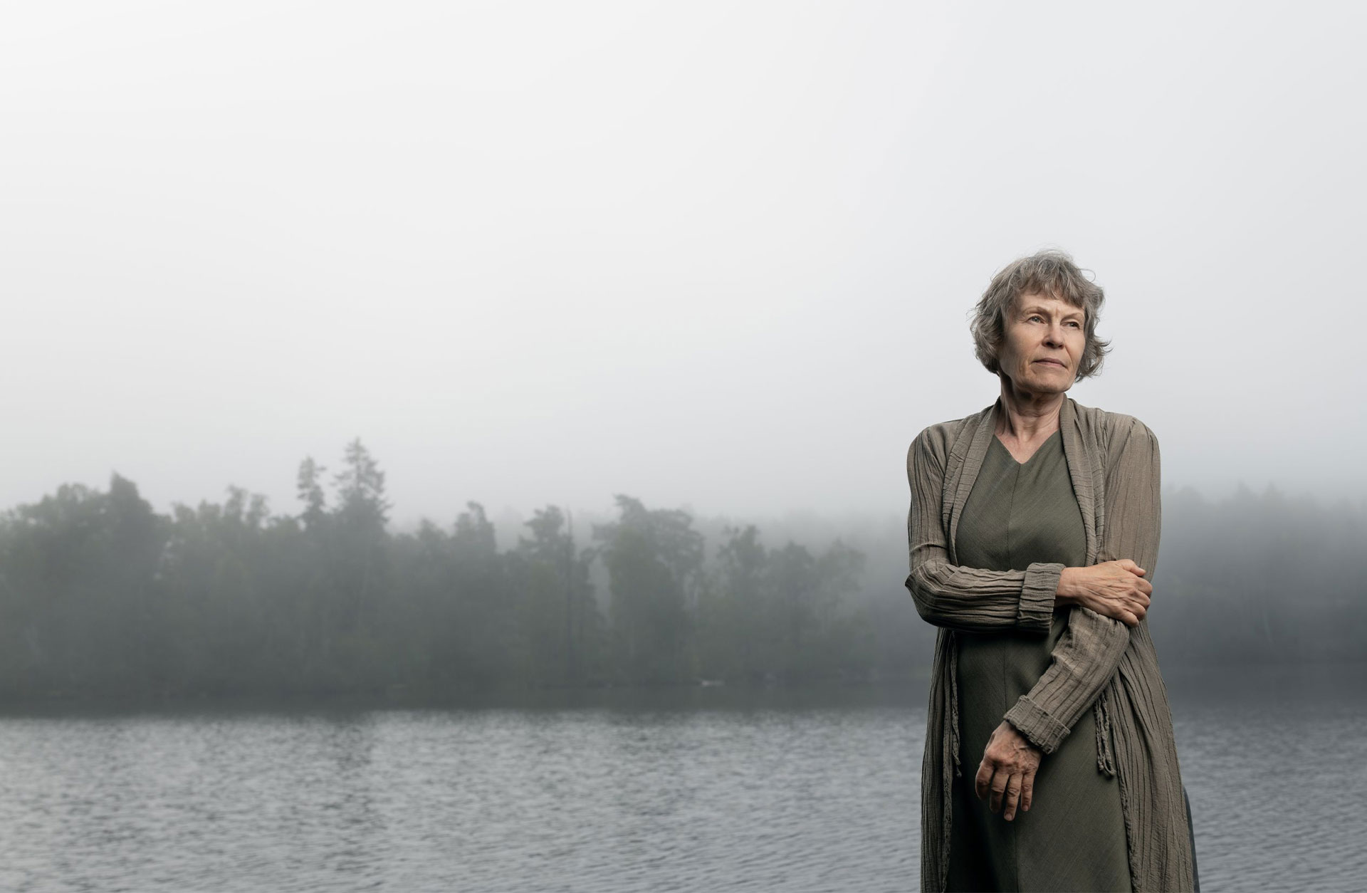 En kvinna i klänning och lång kofta står i skogen vid en sjö. Hon har gråsprängt hår i en kort frisyr och hon tittar förbi kameran.
