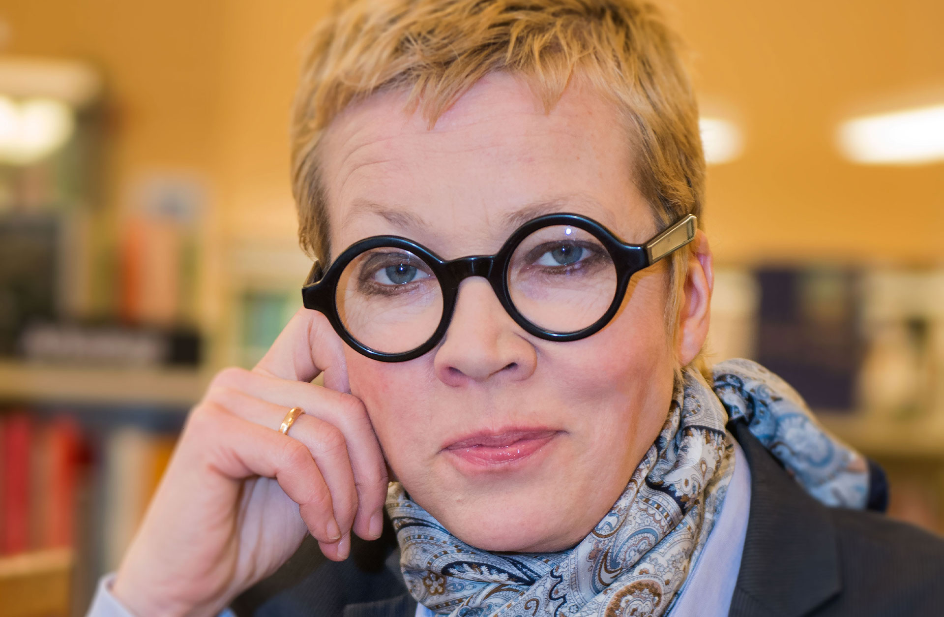 En blond medelålders kvinna med kortklippt frisyr och svarta markanta runda glasögon ler mot kameran.