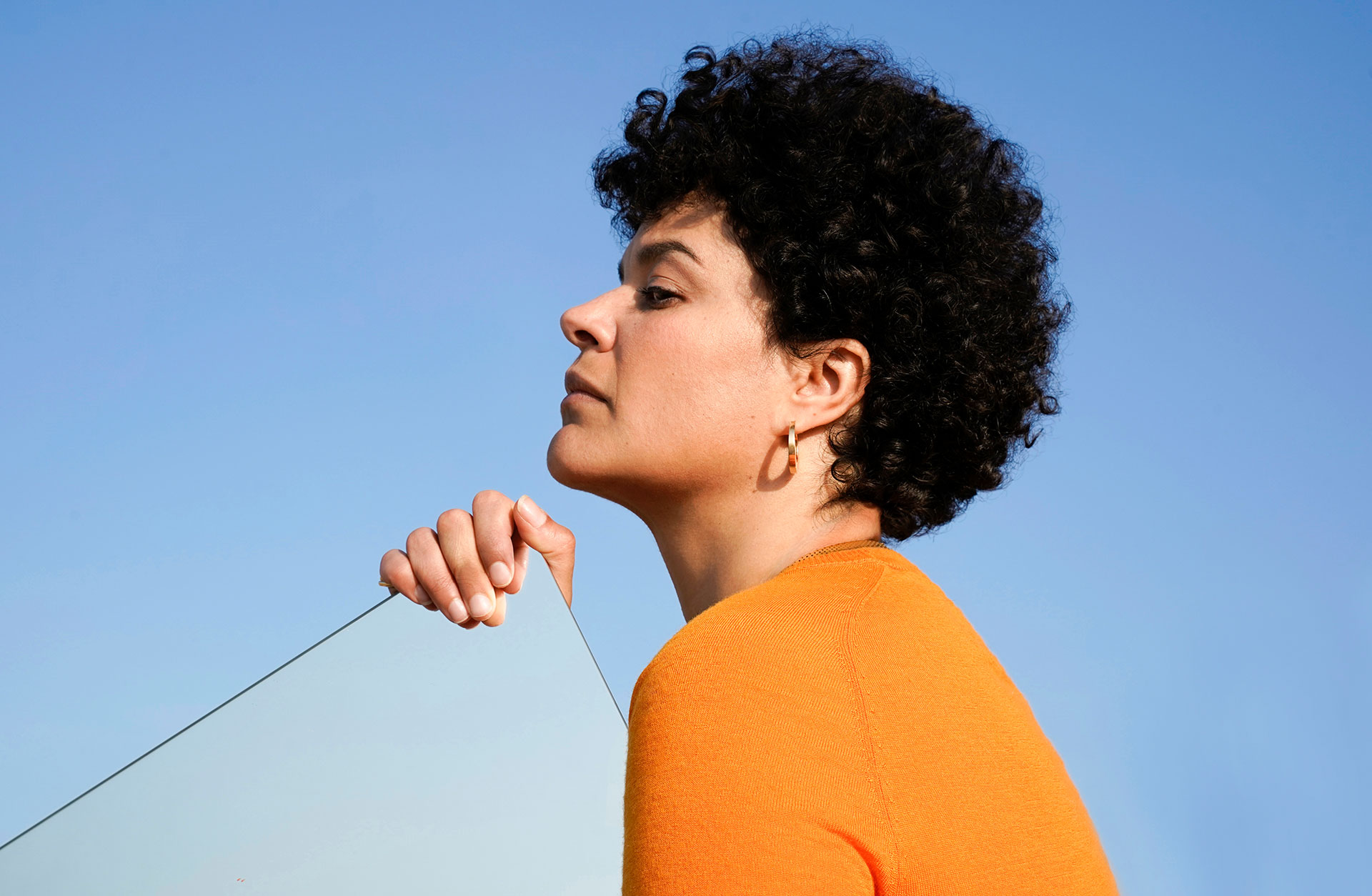 En kvinna i färgstark organge tröja sitter utomhus mot en klarblå himmel. Man ser henne i profil. Hon blundar.