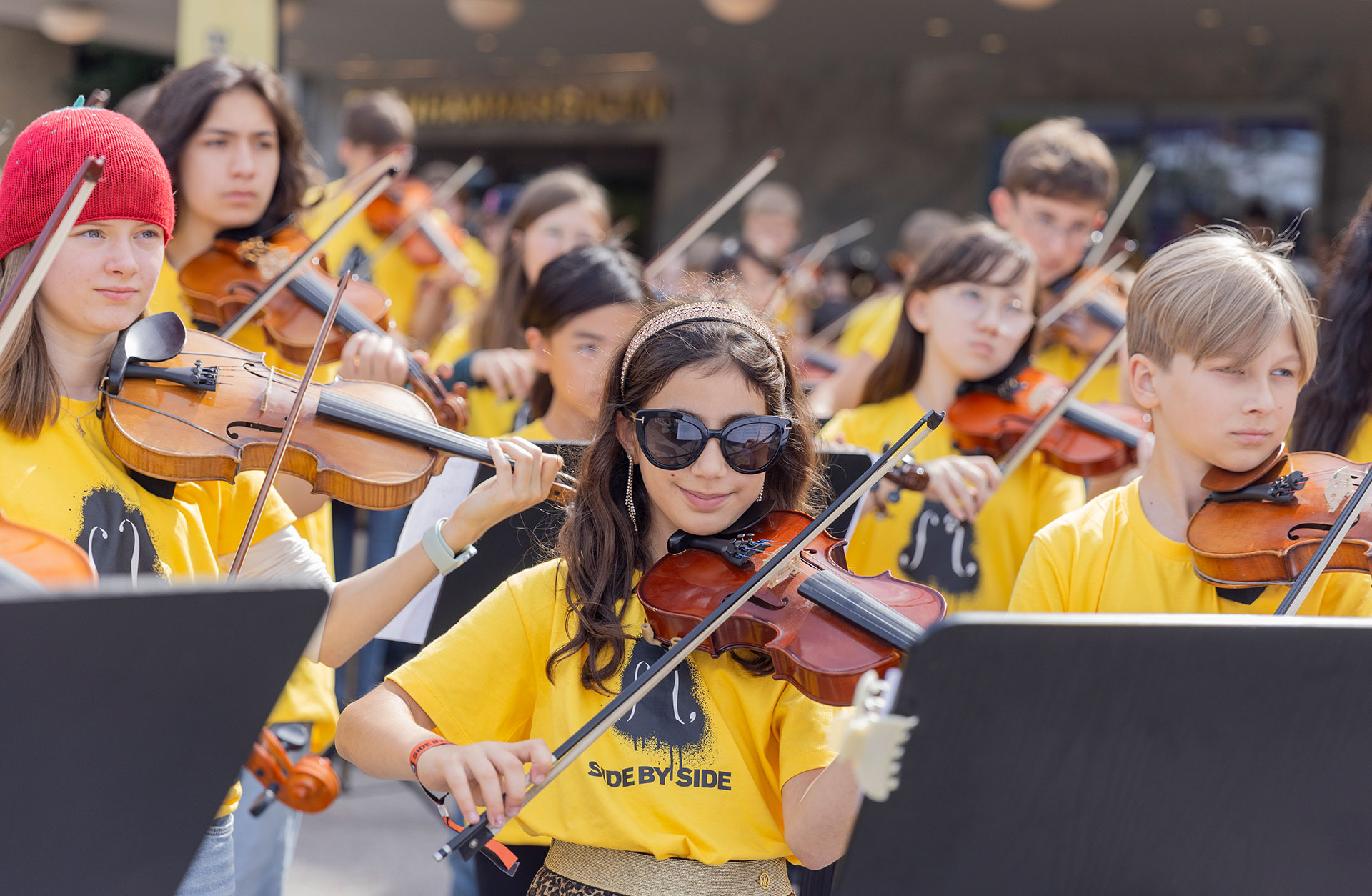 En flicka med långt brunt hår och solglasögon spelar fiol tillsammans med andra barn. Hon tittar rakt in i kameran. De sitter utomhus och spelar i gula t-shirtar.