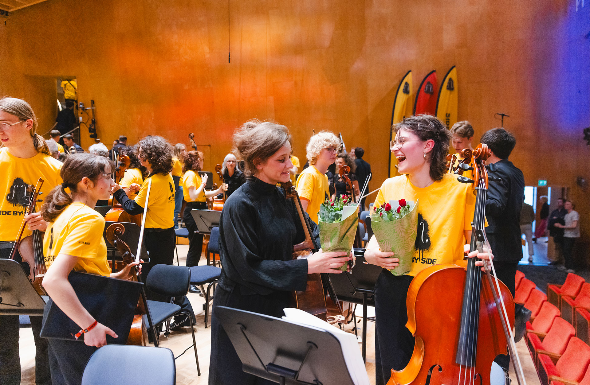 Uppluppen stämning på scenen efter en konsert. Ungdomar i gula t-shirts reser sig. En ung tjej med cello och blommor i handen skrattar och är glad.
