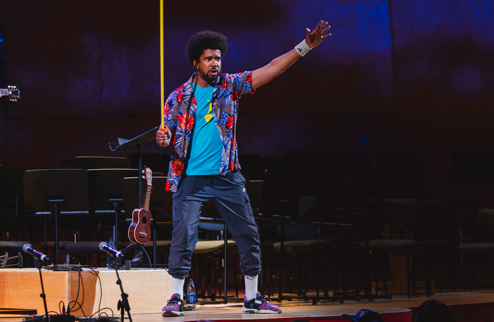 En man står på Konserthusets scen i färd med att berätta något. Han har en blommig skjorta, blå byxor och svart burrigt hår. I handen håller han en gul stav.