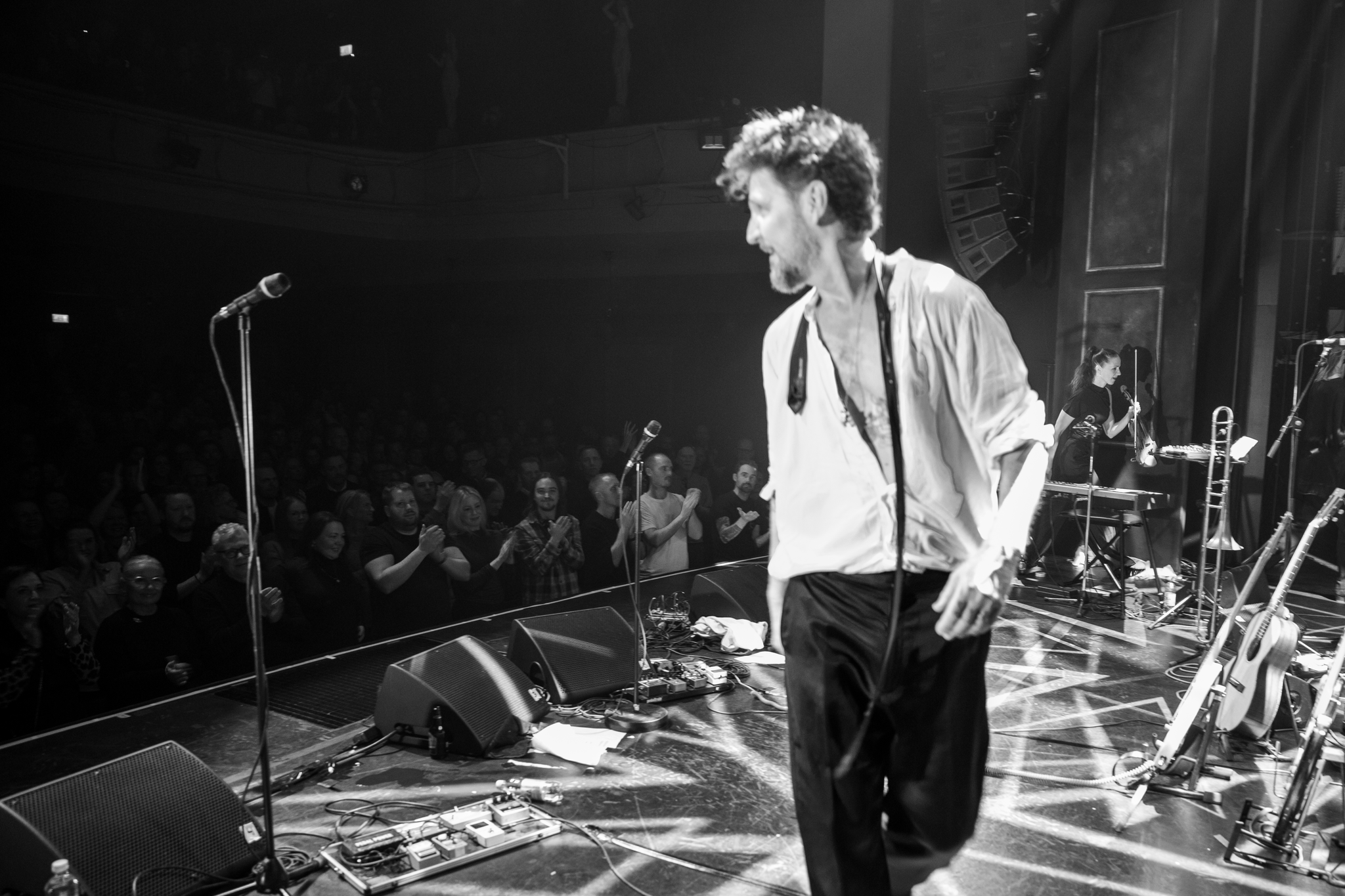 En man i vit skjorta och svarta byxor står på en scen framför publik.