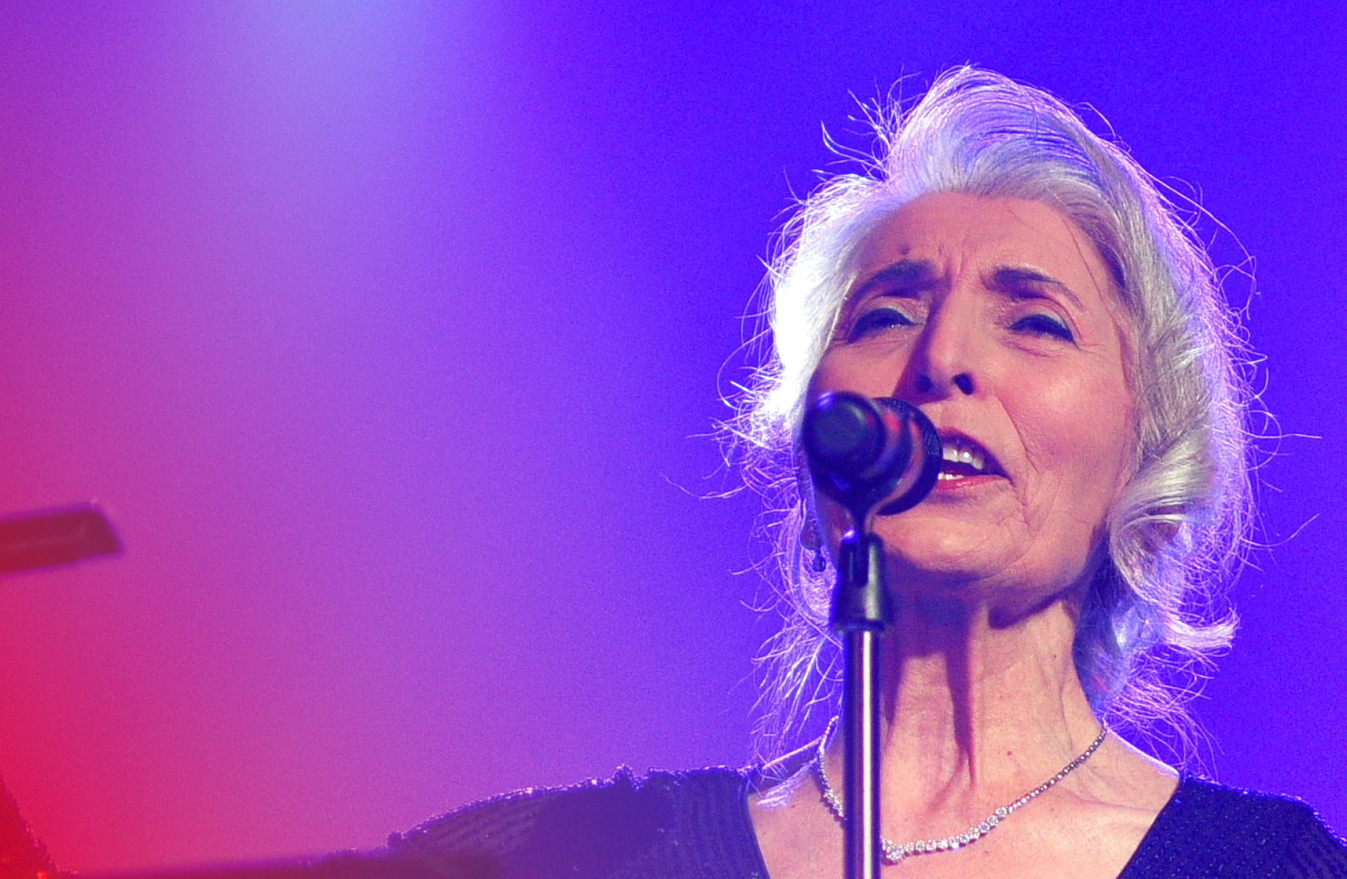 En gråhårig kvinna står framför en violett vägg on sjunger i en mikrofon