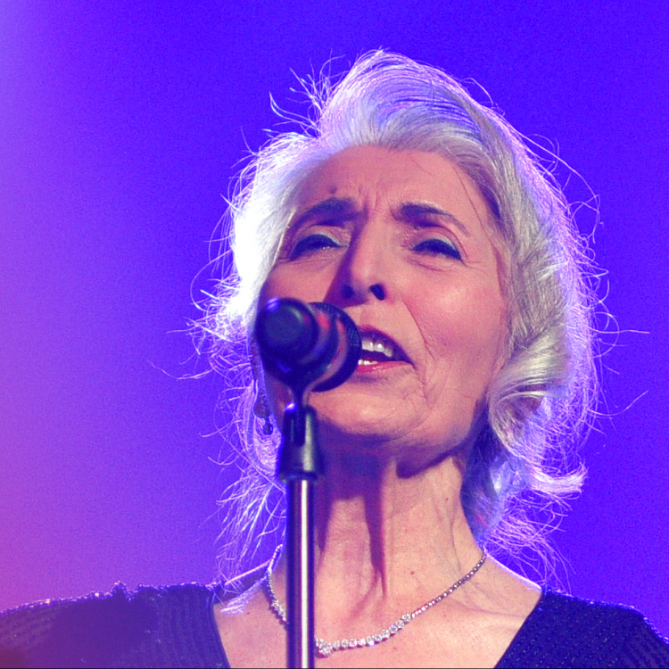 En gråhårig kvinna står framför en violett vägg on sjunger i en mikrofon