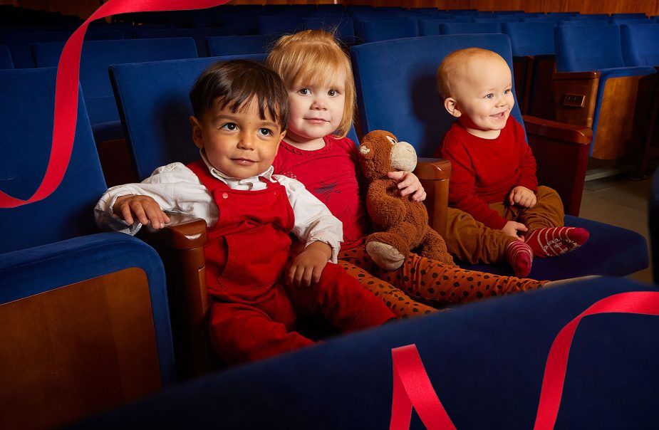 Tre barn sitter i blåa konsertstolar och tittar in i kameran och ler. Det ena barnet håller en nalle i armarna.