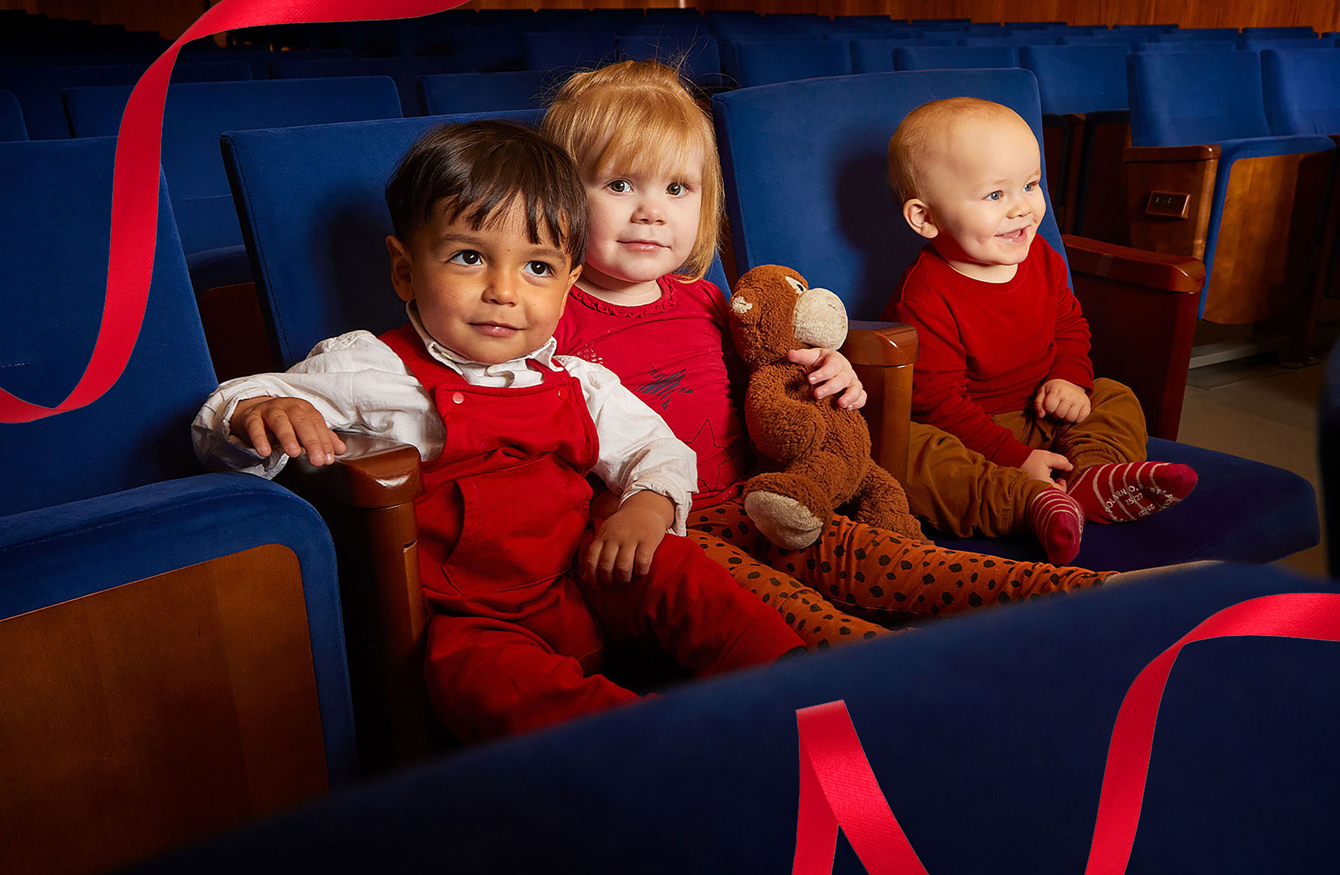 Tre barn sitter i blåa konsertstolar och tittar in i kameran och ler. Det ena barnet håller en nalle i armarna.
