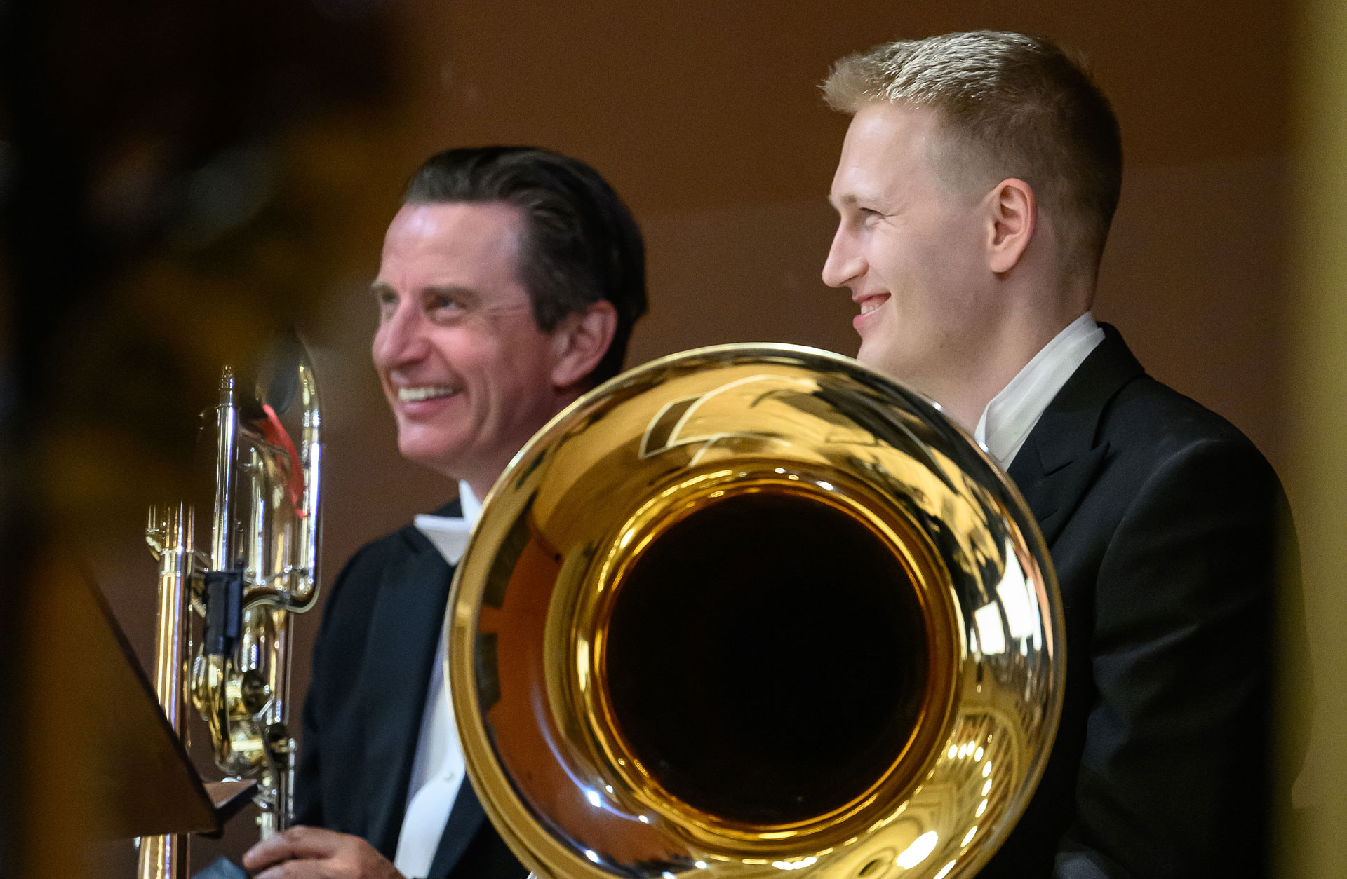 En ung man med sin tuba i knät skrattar. Bredvid sitter en annan man med en trombon.