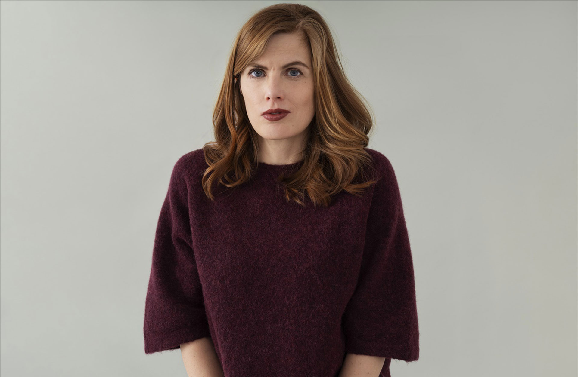 En ung kvinna i stickad tröja och utslaget axellångt hår tittar mot kameran.