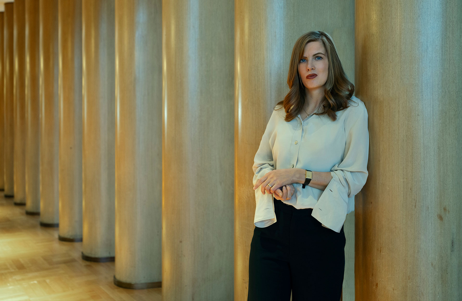 En ung kvinna i vit skjorta står lutad mot en vägg. Hon tittar mot kameran.