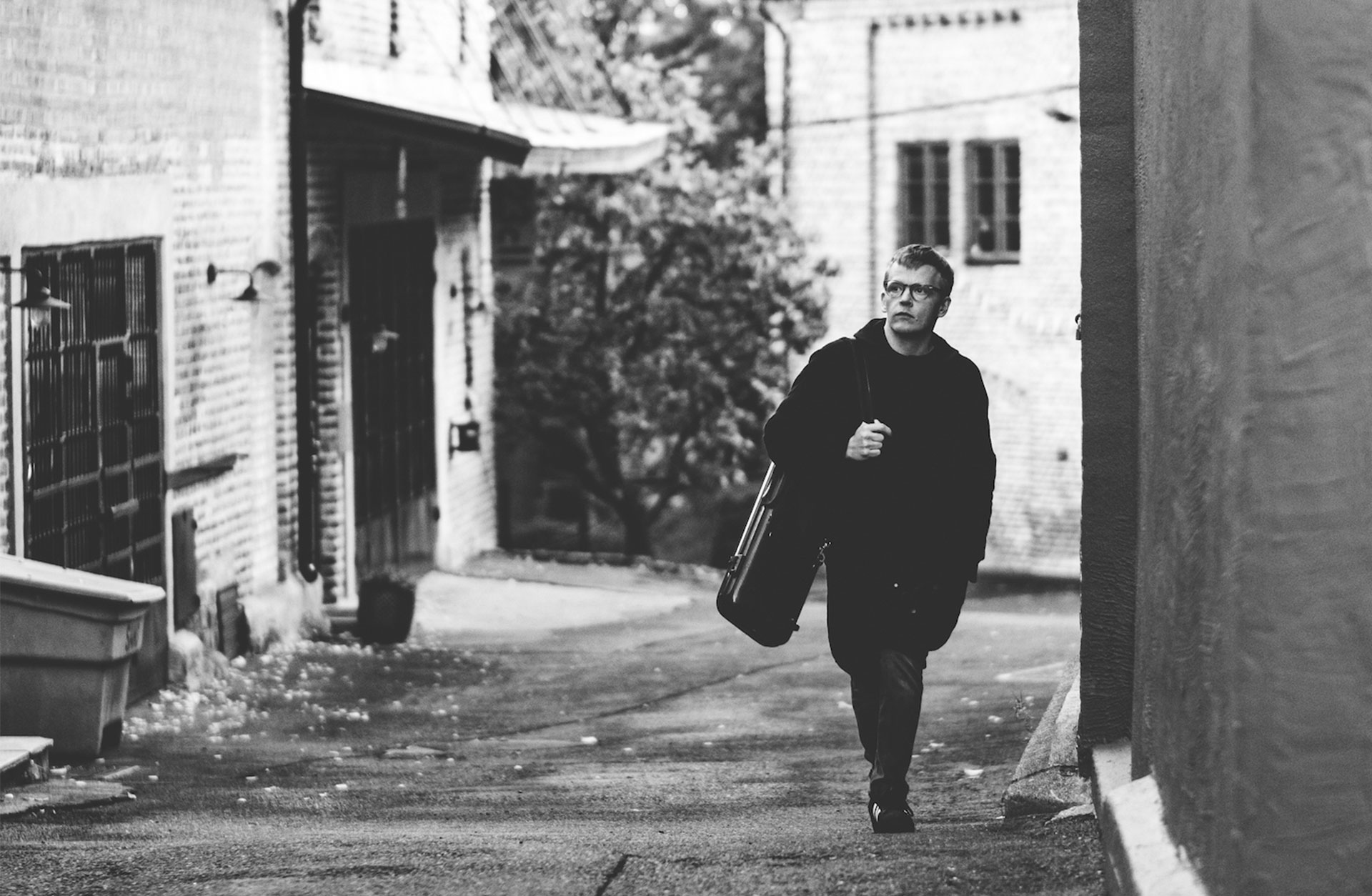 Svartvit bild där en man går på en gata bärandes på ett violinfodral.