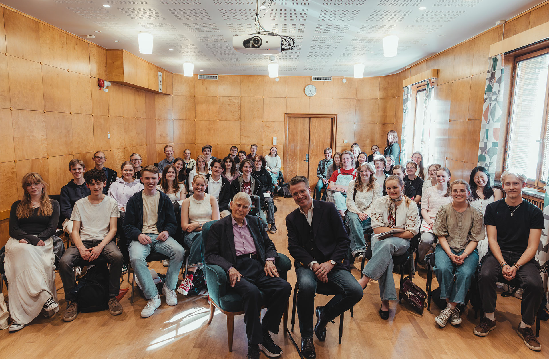 Ett rum med många ungdomar som sitter på stolar bakom två äldre män. alla tittar in i kameran.
