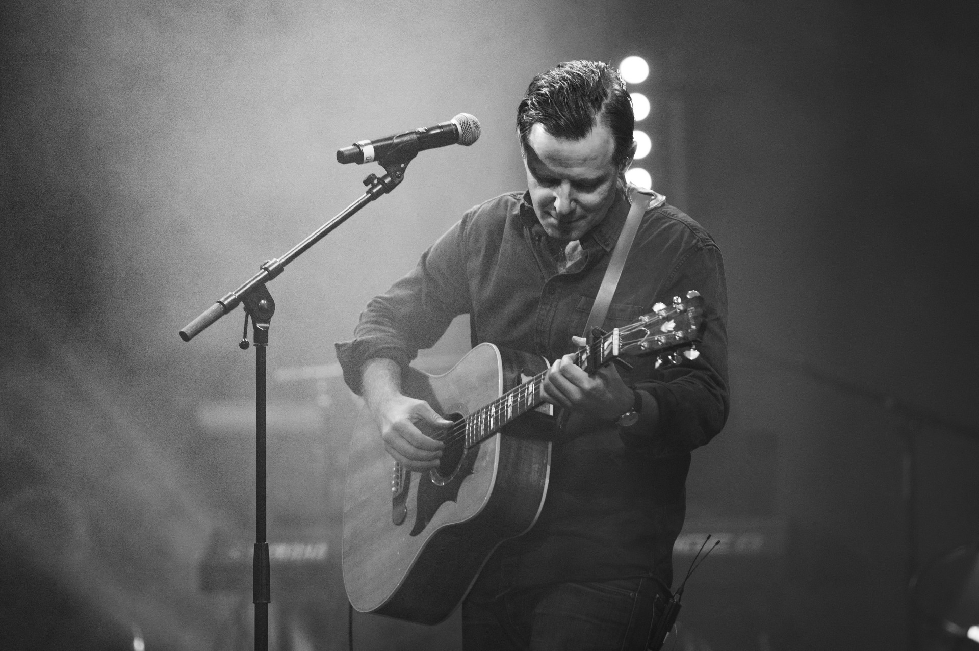 En man står framför en mikrofon och håller i en gitarr. Han har mörka jeans och en örk jeansskjorta på sig