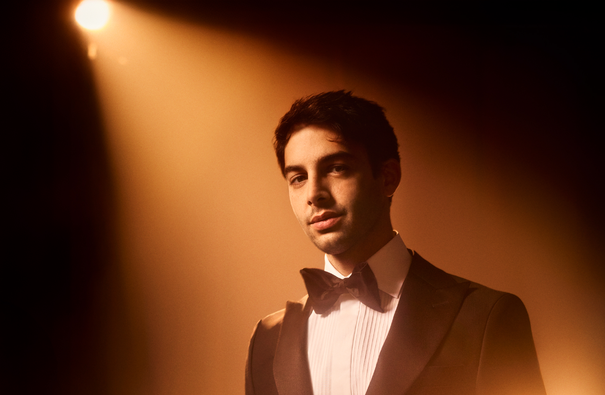 En man i brun kostym, vit skjorta och brun fluga står framför en ljusbrun vägg.