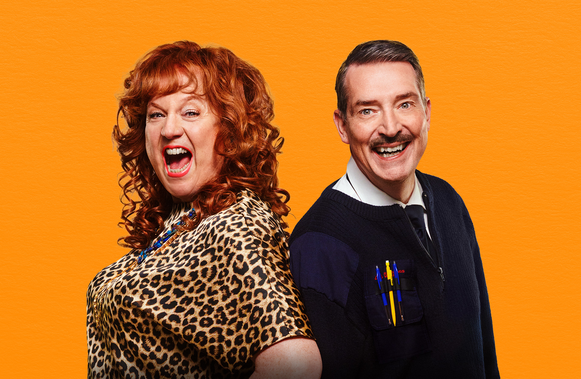 Två män står framför en orange vägg. En av männen är utklädd till kvinna, har rött halvlångt hår och en leopard blu. den andra mannen har mörkt hår, mustas och en mörk tröja.