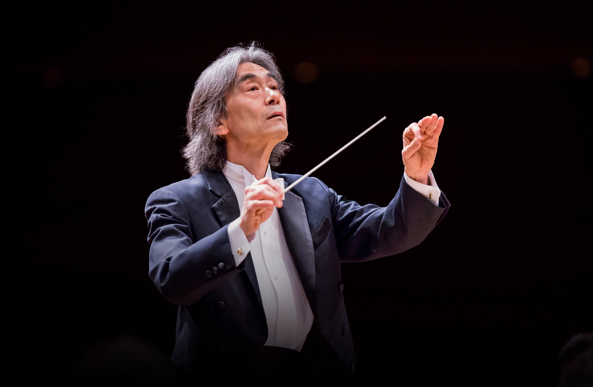 En man med gråsprängt hår och frack som dirigerar med taktpinne.