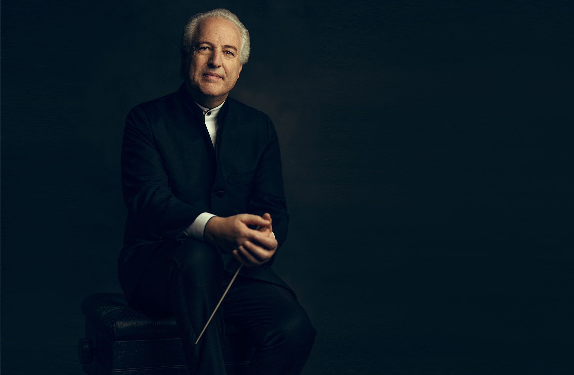 Dirigenten Manfred Honeck sitter på en pianopall i svart kostym, leende med taktpinnen i handen.