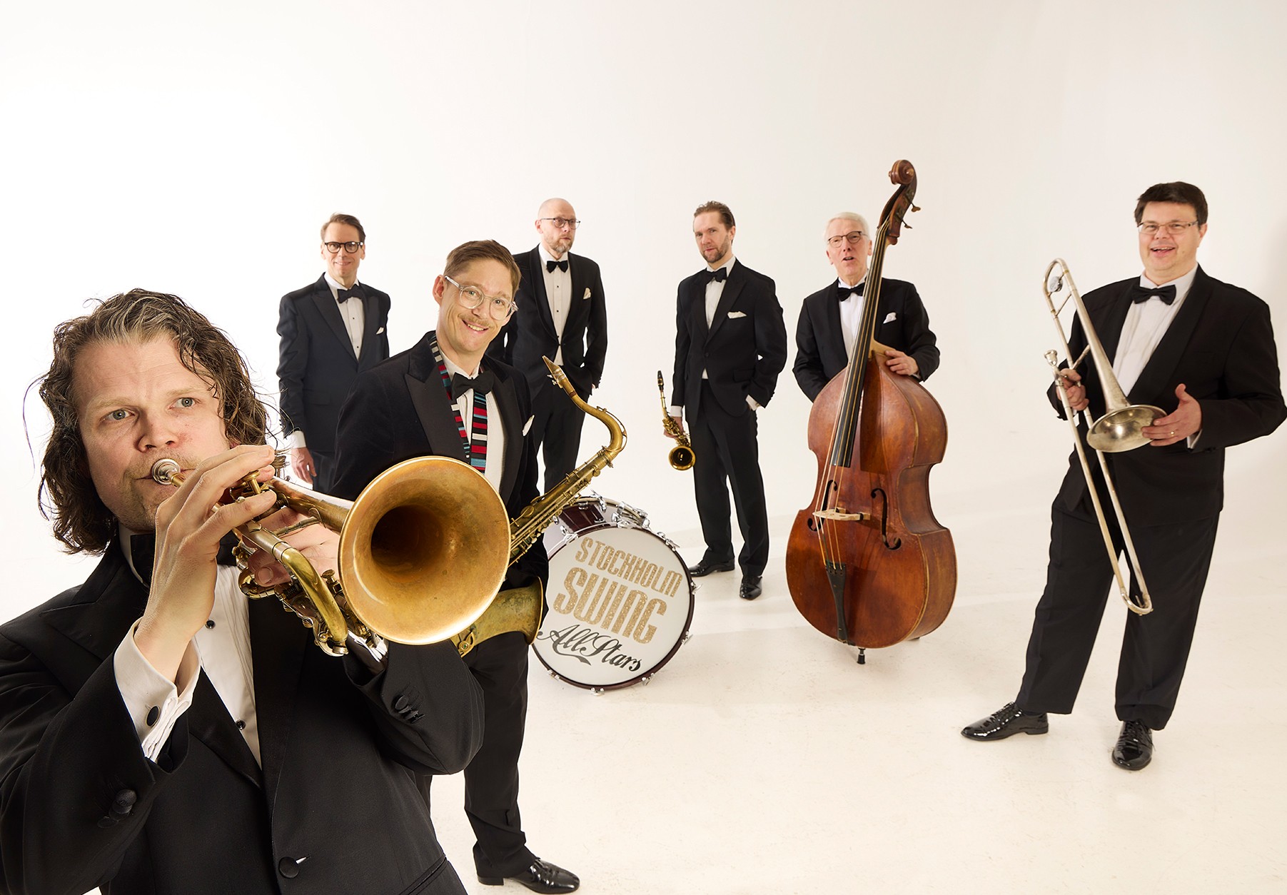 Sju herrar i svart kostym, vit skjorta och svart fluga står framför en vit vägg med olika instrument i händerna.