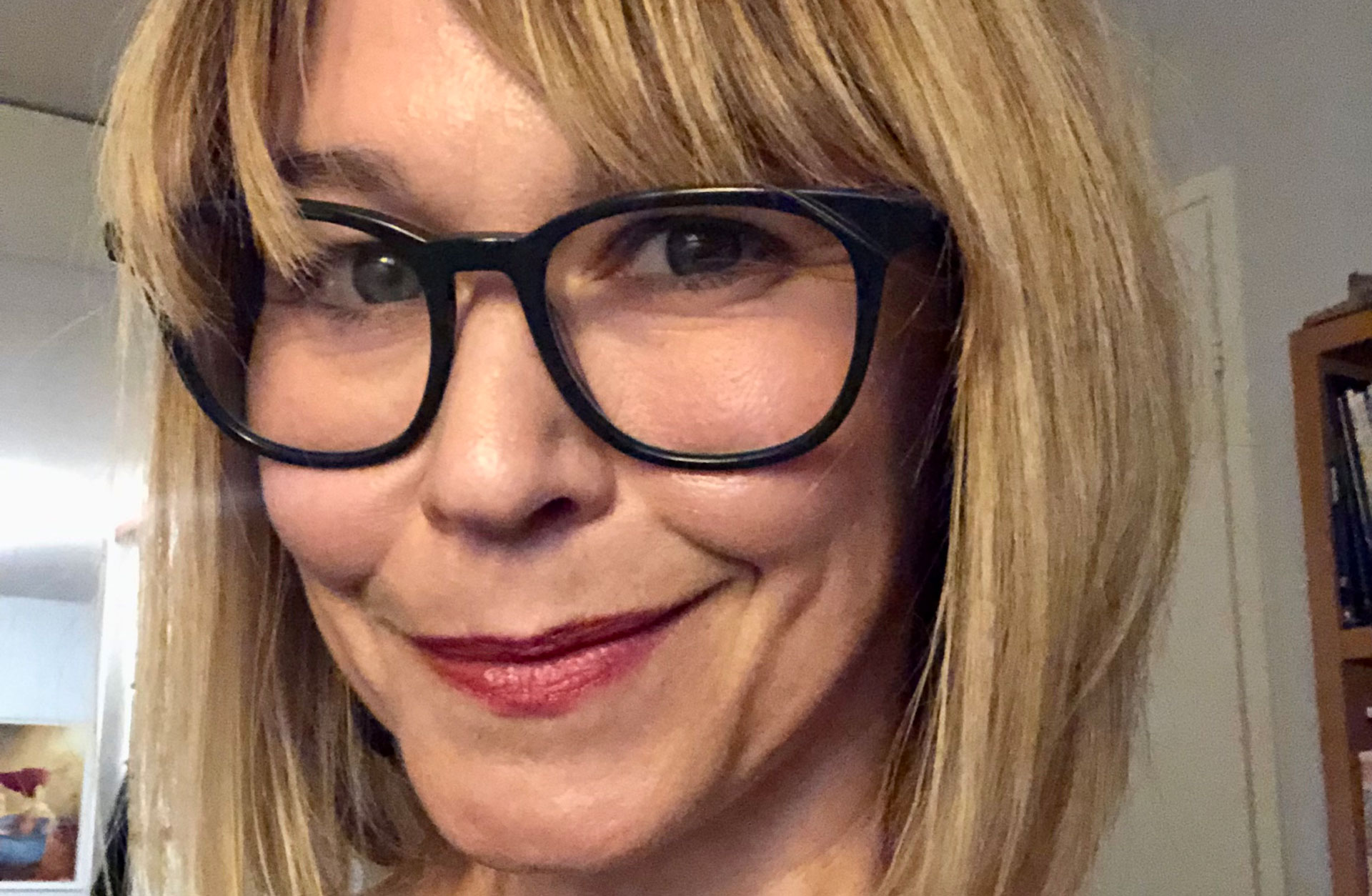 Ansiktsporträtt av en kvinna i blond page och svarta glasögon som ler finurligt.