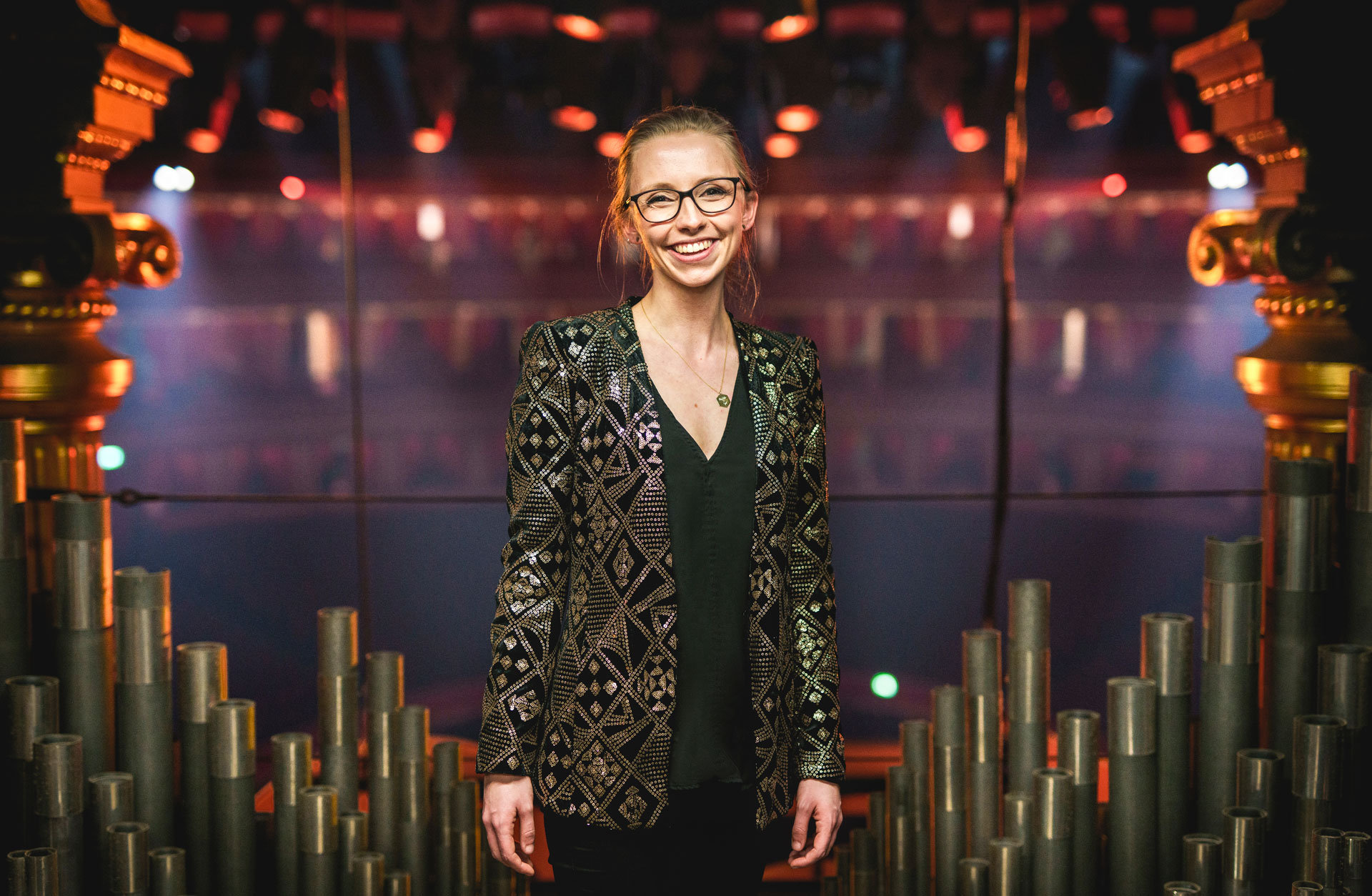 En ung glad kvinna i en guldbrokig kavaj står framför orgelpipor.