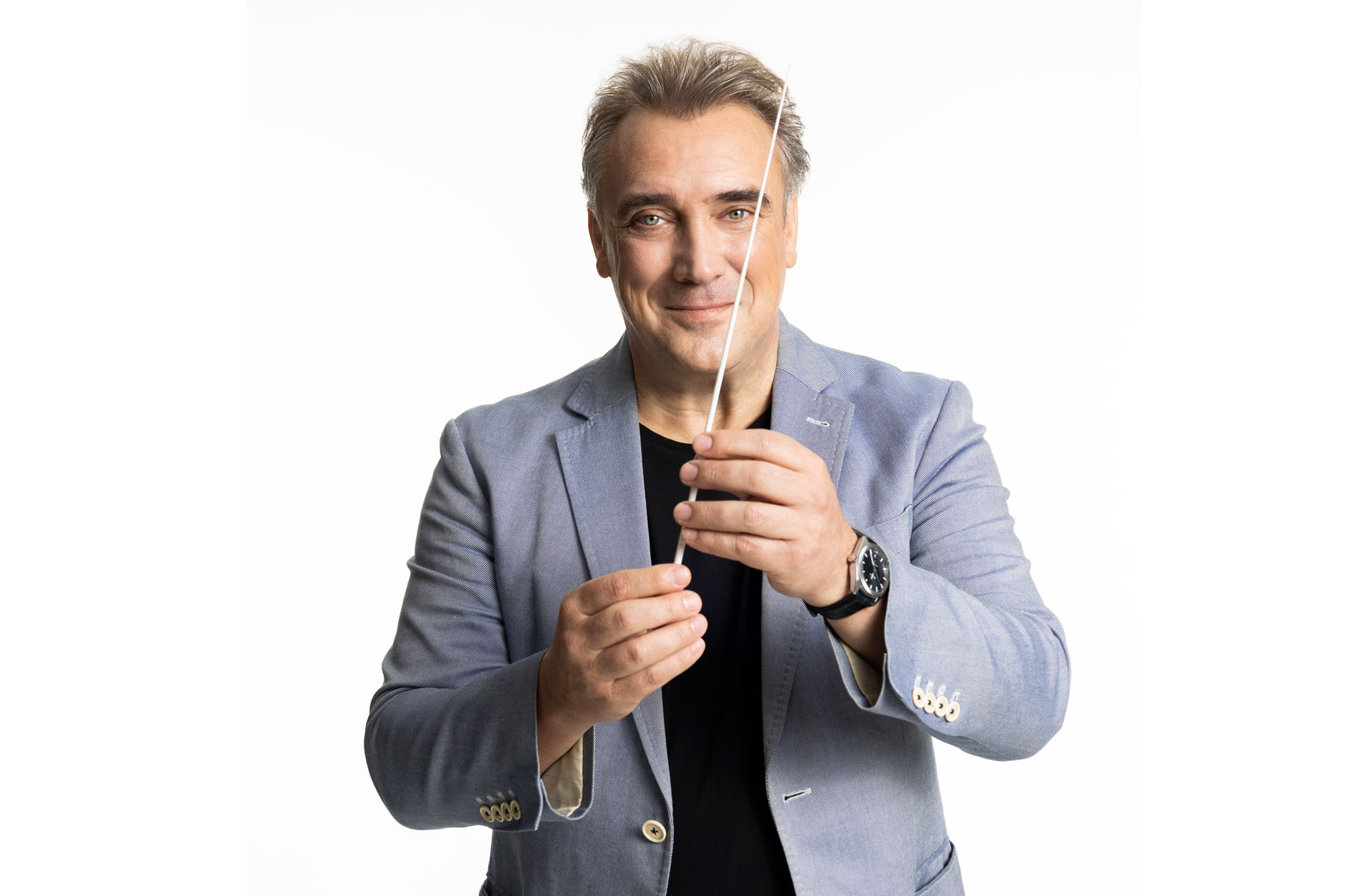 Dirigent fotad mot vit bakgrund håller upp dirigentpinnen mot kameran och ler.