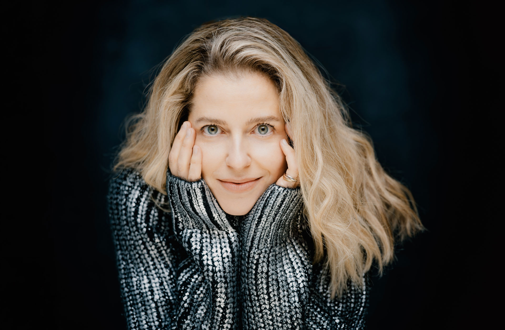 Katarina Canellakis. Bild tagen framifrån. Silverfärgad tröja och huvudet vilandes i händerna.