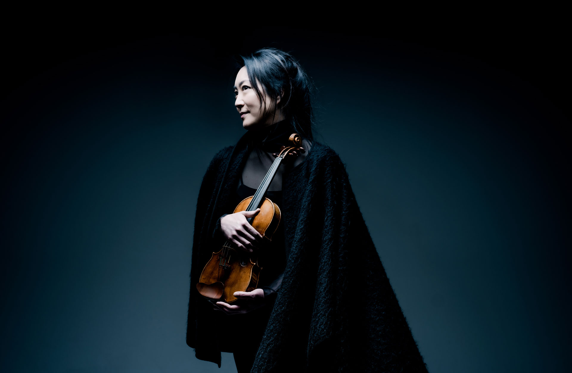 En kvinna med svart långt hår och en svart cape håller i en fiol.
